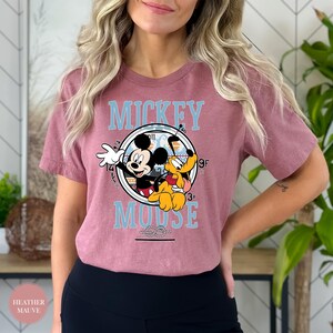 May include: Heather mauve t-shirt featuring a circular graphic with Mickey Mouse and Pluto. The graphic includes the words "Mickey Mouse" in light blue. The shirt is a short-sleeved crew neck.