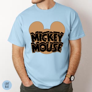 Puede incluir: Camiseta azul claro con un dise&ntilde;o de Mickey Mouse. El dise&ntilde;o incluye un contorno de cabeza de Mickey Mouse de color beige y las palabras "MICKEY MOUSE" en letras negras con un relleno de estampado de leopardo beige. La camiseta es de manga corta.