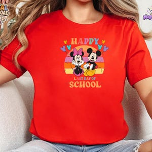 May include: A bright red short-sleeved t-shirt featuring a graphic of Mickey and Minnie Mouse. The design includes a sunset background, colorful balloons, and the text "HAPPY LAST DAY OF SCHOOL" printed in a bold, retro-style font on the front.