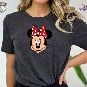 以下が含まれることがあります： ダークグレーの霜降り半袖Tシャツで、前面にミニーマウスのグラフィックがプリントされています。キャラクターは黒い頭、大きな丸い耳を持ち、耳の間の中心には白い水玉模様の鮮やかな赤いリボンが描かれています。