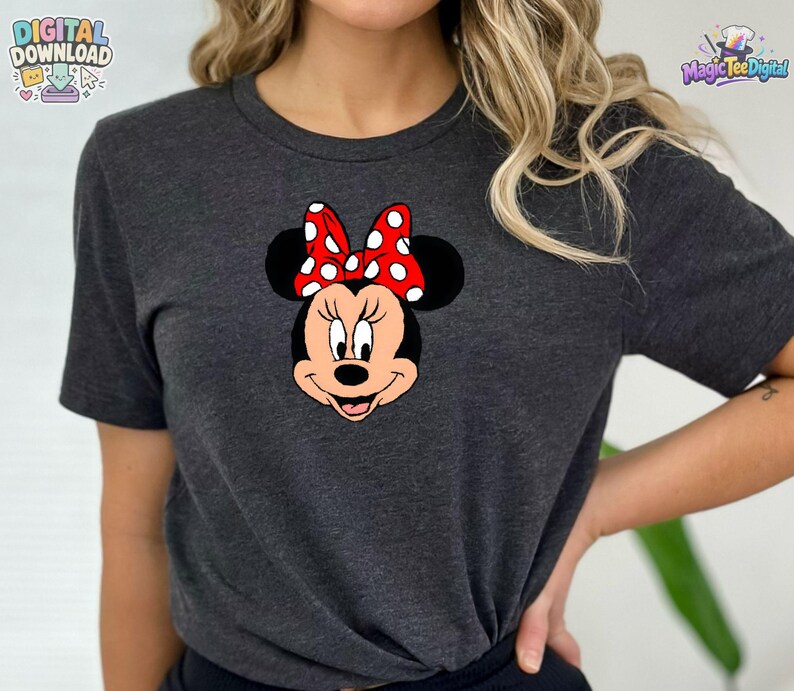 以下が含まれることがあります： ダークグレーの霜降り半袖Tシャツで、前面にミニーマウスのグラフィックがプリントされています。キャラクターは黒い頭、大きな丸い耳を持ち、耳の間の中心には白い水玉模様の鮮やかな赤いリボンが描かれています。