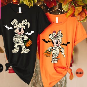 May include: Black and orange Halloween t-shirts featuring Mickey and Minnie Mouse dressed as mummies, holding jack-o'-lanterns. White bats are printed above the characters. The text "Ship Next Day" is visible on the left side of the image.