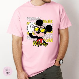 May include: A light pink t-shirt featuring a graphic of Mickey Mouse wearing sunglasses. The design includes the word "Mickey" in yellow and the word "MOUSE" repeated in the background. The shirt is a soft pink color.