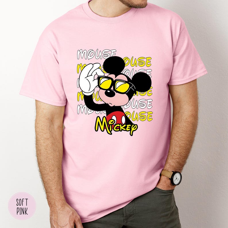 May include: A light pink t-shirt featuring a graphic of Mickey Mouse wearing sunglasses. The design includes the word "Mickey" in yellow and the word "MOUSE" repeated in the background. The shirt is a soft pink color.