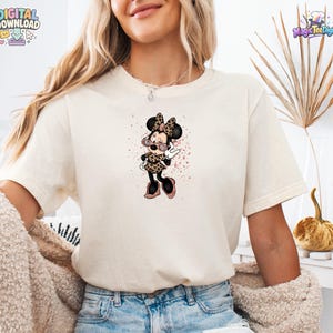 May include: A cream-coloured short-sleeve t-shirt featuring a graphic of a cartoon mouse character wearing leopard print clothing and pink-tinted sunglasses. The character is surrounded by pink glittery splatters on the front of the shirt.