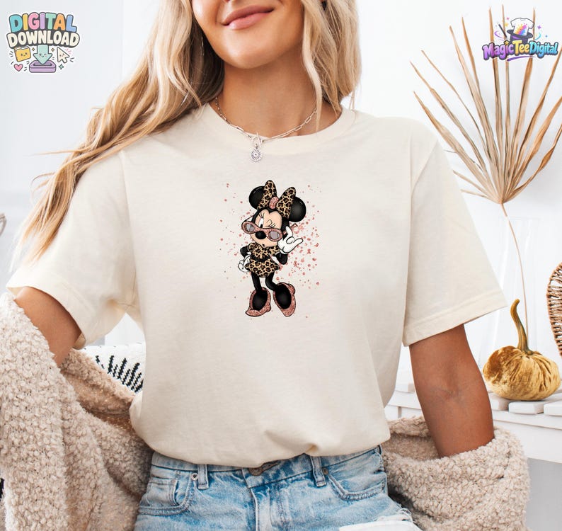 May include: A cream-coloured short-sleeve t-shirt featuring a graphic of a cartoon mouse character wearing leopard print clothing and pink-tinted sunglasses. The character is surrounded by pink glittery splatters on the front of the shirt.
