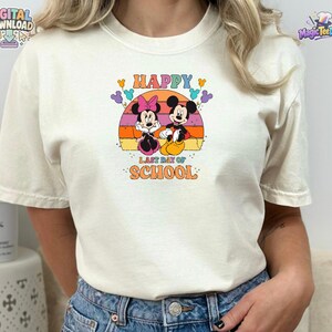 May include: A cream-colored short-sleeve t-shirt featuring a graphic of Mickey and Minnie Mouse. The design includes a sunset background, colorful balloons, and the text "HAPPY LAST DAY OF SCHOOL" in a retro-style font centered on the chest.