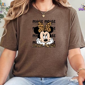 Pode incluir: Uma t-shirt castanha de manga curta com um gr&aacute;fico da Minnie Mouse com um la&ccedil;o de estampado de leopardo. O texto "MINNIE MOUSE" &eacute; repetido no fundo e impresso com um padr&atilde;o de leopardo na parte inferior do design gr&aacute;fico frontal.