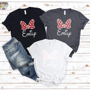 May include: Three t-shirts with a Minnie Mouse bow design and the name "Emily" in a script font. The shirts are navy, gray, and white. A pair of blue jeans is also in the image. The bow on the navy and gray shirts is red with white polka dots.