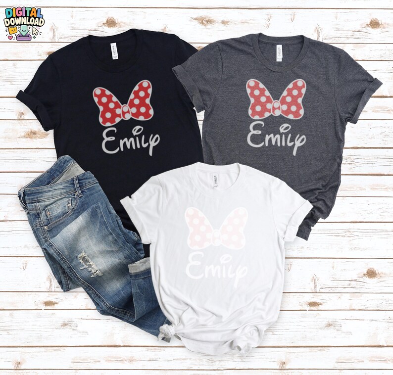 May include: Three t-shirts with a Minnie Mouse bow design and the name "Emily" in a script font. The shirts are navy, gray, and white. A pair of blue jeans is also in the image. The bow on the navy and gray shirts is red with white polka dots.