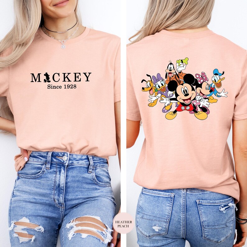 Puede incluir: Camiseta color melocot&oacute;n con el texto "MICKEY Since 1928" en la parte delantera. La parte trasera de la camiseta muestra un gr&aacute;fico colorido de Mickey Mouse y sus amigos. La camiseta se combina con vaqueros azules.