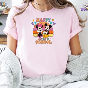 May include: A light pink t-shirt featuring a graphic of Minnie and Mickey Mouse sitting in front of a sunset. The text reads "HAPPY LAST DAY OF SCHOOL" in orange and blue letters, surrounded by small colorful Mickey-shaped balloon icons.