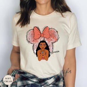 May include: Cream-colored t-shirt featuring a graphic of a cartoon character with a bow and the word "Moana". The bow is coral with white polka dots. The shirt has the words "Soft Cream" on the bottom left.