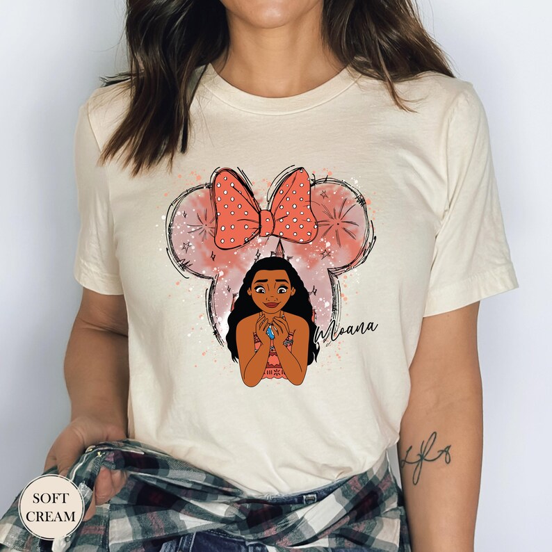 May include: Cream-colored t-shirt featuring a graphic of a cartoon character with a bow and the word "Moana". The bow is coral with white polka dots. The shirt has the words "Soft Cream" on the bottom left.