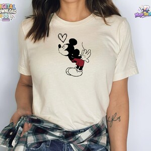 May include: A cream-colored short-sleeve t-shirt featuring a black and red graphic of a cartoon mouse blowing a kiss with a small heart outline above its head. The shirt is worn with a dark green, black, and white plaid flannel shirt tied around the waist.