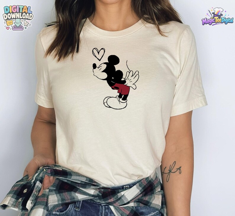 May include: A cream-colored short-sleeve t-shirt featuring a black and red graphic of a cartoon mouse blowing a kiss with a small heart outline above its head. The shirt is worn with a dark green, black, and white plaid flannel shirt tied around the waist.