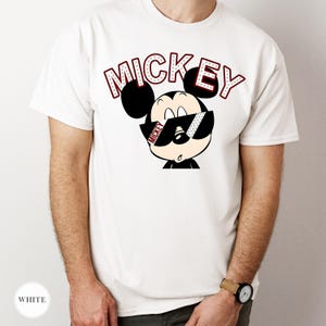 May include: White t-shirt featuring a cartoon Mickey Mouse wearing sunglasses. The word "MICKEY" is printed above the character in a red and white font. The shirt is a classic crew neck style.