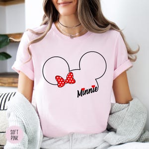 May include: A light pink t-shirt featuring a black outline of Mickey Mouse ears with a red polka dot bow. The word "Minnie" is written in black script below the bow, with two small red hearts. The shirt also has the words "Soft Pink" on the bottom left.