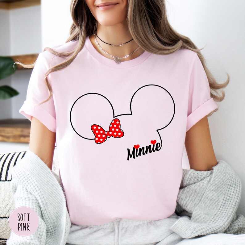 May include: A light pink t-shirt featuring a black outline of Mickey Mouse ears with a red polka dot bow. The word "Minnie" is written in black script below the bow, with two small red hearts. The shirt also has the words "Soft Pink" on the bottom left.