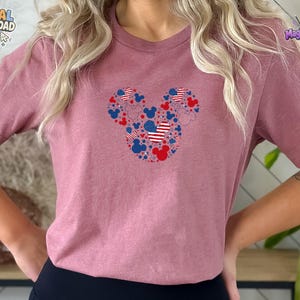 Puede incluir: Una camiseta de manga corta color malva con un gr&aacute;fico central de una gran silueta de cabeza de Mickey Mouse compuesta por iconos m&aacute;s peque&ntilde;os de cabezas de Mickey, estrellas y patrones de bandera estadounidense en rojo, blanco y azul.