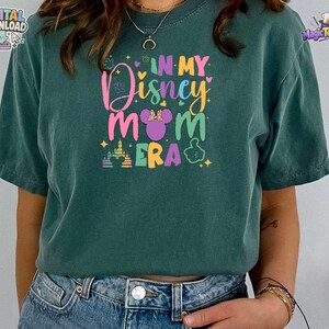 May include: A dark green short-sleeve t-shirt featuring a colorful graphic print that reads IN MY Disney MOM ERA. The design includes a purple mouse ear silhouette with a bow, a castle outline, and small star accents in pink, yellow, and blue.