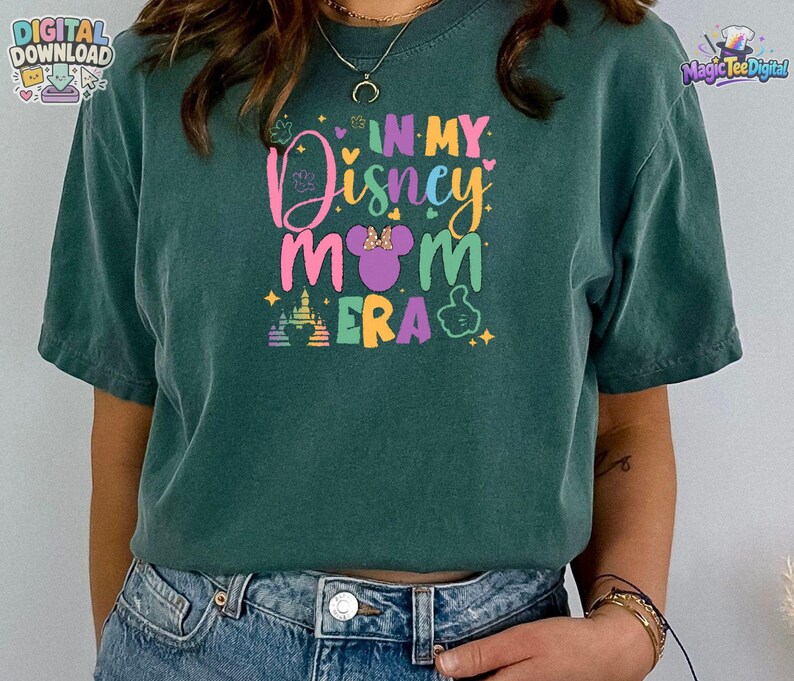 May include: A dark green short-sleeve t-shirt featuring a colorful graphic print that reads IN MY Disney MOM ERA. The design includes a purple mouse ear silhouette with a bow, a castle outline, and small star accents in pink, yellow, and blue.