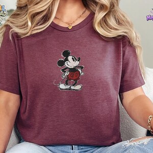 May include: A maroon short-sleeved t-shirt featuring a centered graphic of a vintage-style Mickey Mouse character. The character is depicted in black, white, and red with a distressed texture. The shirt has a crew neckline and a heathered fabric finish.