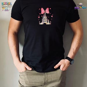 Pu&ograve; includere: Una t-shirt nera a maniche corte con una grafica centrale che mostra la sagoma di un castello sormontata da un fiocco rosa e orecchie da topo, circondata da piccoli cuori rosa. Il design utilizza uno stile acquerello con toni rosa e bianchi.