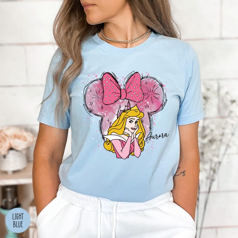 May include: Pale blue t-shirt with a pink bow and Aurora cartoon graphic. The design includes a pink and white Mickey Mouse head outline and the name "Aurora" in cursive.