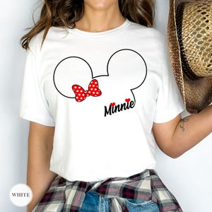 May include: White t-shirt featuring a Minnie Mouse design. The design includes a black outline of Minnie's head, a red polka-dot bow, the name "Minnie" in black script, and two red hearts. The shirt is short-sleeved.