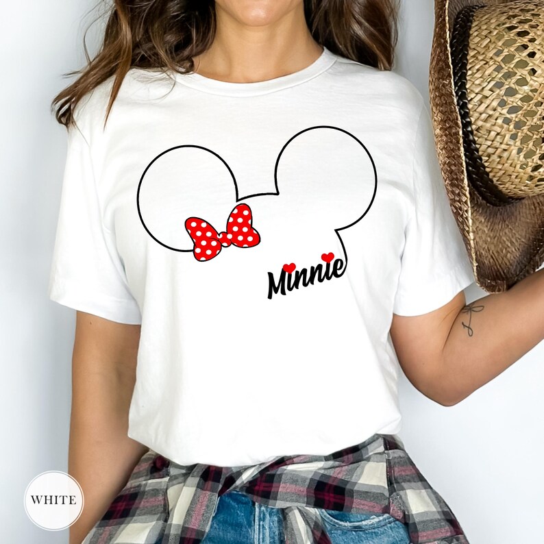 May include: White t-shirt featuring a Minnie Mouse design. The design includes a black outline of Minnie's head, a red polka-dot bow, the name "Minnie" in black script, and two red hearts. The shirt is short-sleeved.