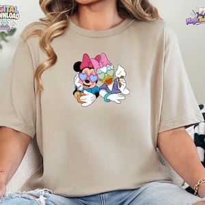 May include: A beige short-sleeved t-shirt featuring a graphic print of Minnie Mouse and Daisy Duck wearing heart-shaped sunglasses. Minnie holds an orange drink with a straw, while Daisy holds a white ice cream cone. The design is centered.