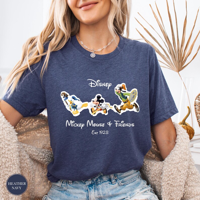 May include: Heather navy blue t-shirt featuring the Disney logo and the phrase "Mickey Mouse & Friends Est 1928." The shirt displays cartoon images of Donald Duck, Mickey Mouse, and Goofy. The shirt is a short-sleeved crew neck.