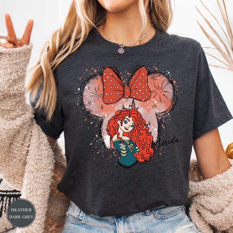Puede incluir: Camiseta gris oscuro con una ilustraci&oacute;n de dibujos animados de una princesa pelirroja, un lazo y el nombre "Merida". El dise&ntilde;o est&aacute; dentro de un contorno en forma de Mickey Mouse con un lazo de lunares.