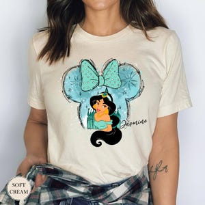 Puede incluir: Camiseta color crema con un gr&aacute;fico de la Princesa Jasmine dentro de un contorno de orejas de Mickey Mouse. El dise&ntilde;o incluye un lazo turquesa, estrellas y el nombre "Jasmine". La camiseta tiene cuello redondo.