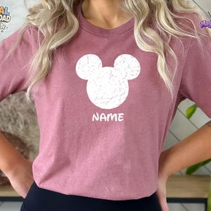 May include: A mauve short-sleeved t-shirt featuring a white distressed graphic of a mouse ear silhouette centered on the chest. Below the graphic, the word NAME is printed in white capital letters. The shirt has a crew neckline and a plain back.