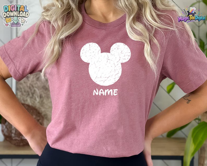 May include: A mauve short-sleeved t-shirt featuring a white distressed graphic of a mouse ear silhouette centered on the chest. Below the graphic, the word NAME is printed in white capital letters. The shirt has a crew neckline and a plain back.
