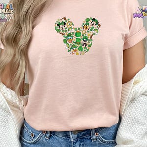 Pu&ograve; includere: Una t-shirt a maniche corte color rosa chiaro con una grafica centrale a forma di silhouette di testa di topo. Il disegno &egrave; composto da vari personaggi dei cartoni animati con cappelli verdi, trifogli, monete d'oro e pentole d'oro.