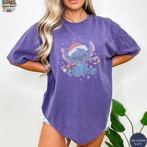May include: Heather navy blue t-shirt featuring a cartoon character wearing a Santa hat and a scarf with Christmas lights. The graphic is in shades of blue, red, and white. The shirt has short sleeves and a relaxed fit.