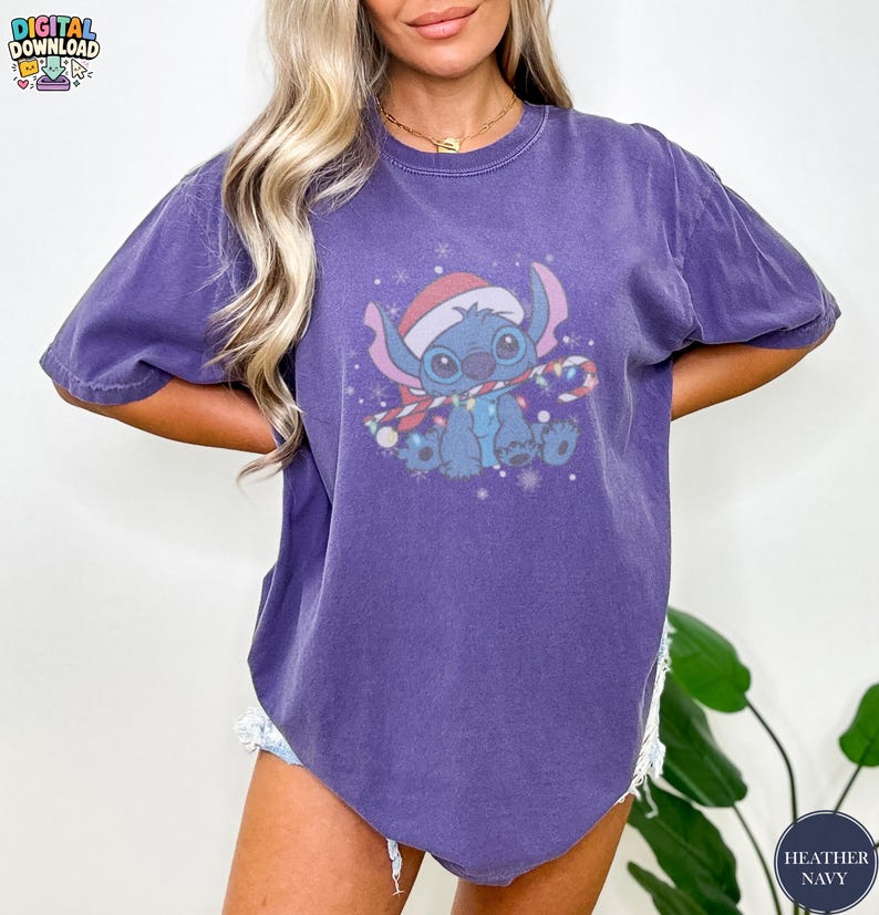 May include: Heather navy blue t-shirt featuring a cartoon character wearing a Santa hat and a scarf with Christmas lights. The graphic is in shades of blue, red, and white. The shirt has short sleeves and a relaxed fit.