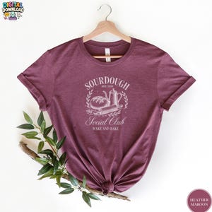 May include: A heather maroon short-sleeve t-shirt featuring a white graphic print of a loaf of bread, a pitcher, and a rolling pin. The text reads SOURDOUGH EST. 2020 SOCIAL CLUB WAKE AND BAKE. The shirt is displayed on a wooden hanger.