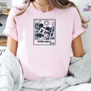 K&ouml;nnte beinhalten: Ein hellrosa T-Shirt mit einem schwarzen Grafikdruck eines quadratischen Rahmens im Polaroid-Stil, der Zeichentrickfiguren wie Mickey Maus, Minnie Maus, Donald Duck und Goofy enth&auml;lt, mit dem Text Disney World 2024 unter dem Rahmen.