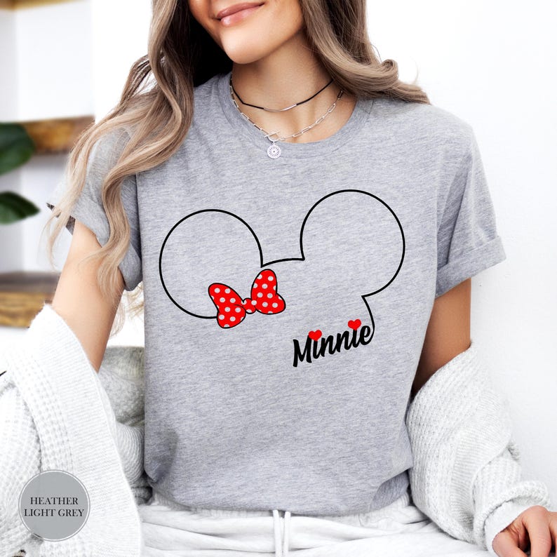 May include: Heather light grey t-shirt featuring a Minnie Mouse design. The design includes black Mickey Mouse ears, a red polka dot bow, and the name "Minnie" in black script with two red hearts. The shirt is a casual style.