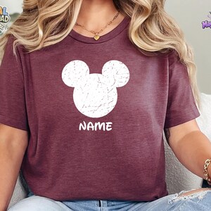 May include: A maroon short-sleeved t-shirt featuring a white distressed graphic of a mouse head silhouette centered on the chest. Below the mouse head graphic, the word NAME is printed in a bold, white, sans-serif font on the front of the shirt.
