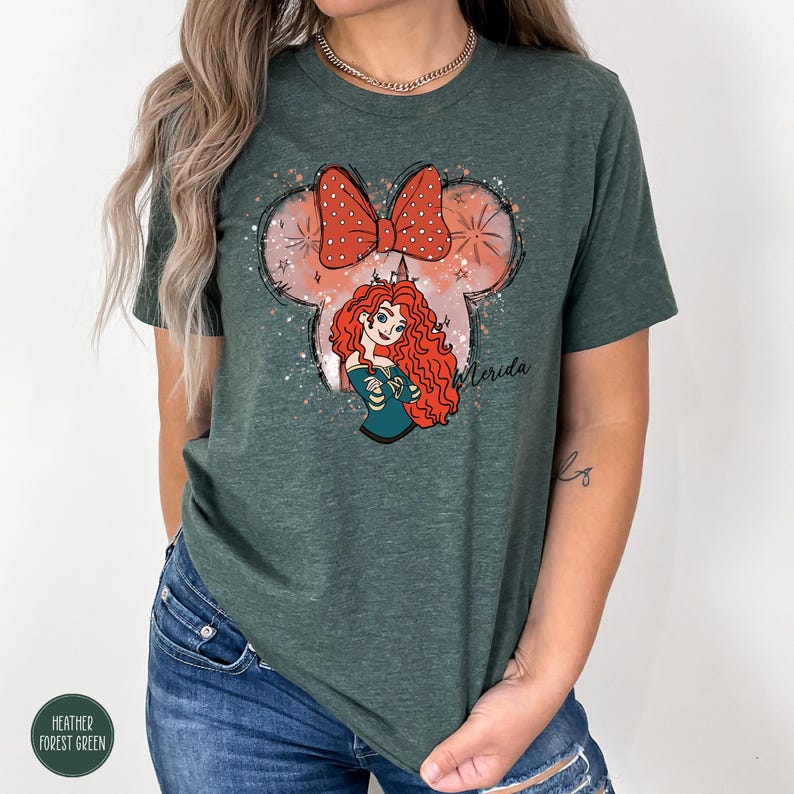 Puede incluir: Camiseta verde bosque jaspeado con una ilustraci&oacute;n de dibujos animados de Merida de Brave. El dise&ntilde;o incluye un lazo rojo con lunares blancos, una silueta de orejas de Mickey Mouse y el nombre "Merida" en cursiva. La camiseta tiene cuello redondo.