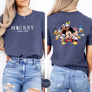 Puede incluir: Camiseta azul marino jaspeada con el texto "Mickey Since 1928" en la parte delantera. La parte trasera de la camiseta muestra un gr&aacute;fico colorido de Mickey Mouse y sus amigos. La camiseta se combina con vaqueros azules.