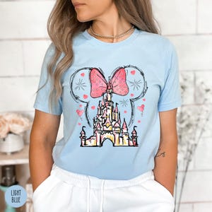 Puede incluir: Camiseta azul claro con un dise&ntilde;o de castillo, un lazo rosa y detalles de coraz&oacute;n. El dise&ntilde;o est&aacute; enmarcado por un contorno de oreja de Mickey Mouse. Las palabras "LIGHT BLUE" est&aacute;n impresas en la parte inferior izquierda de la imagen.