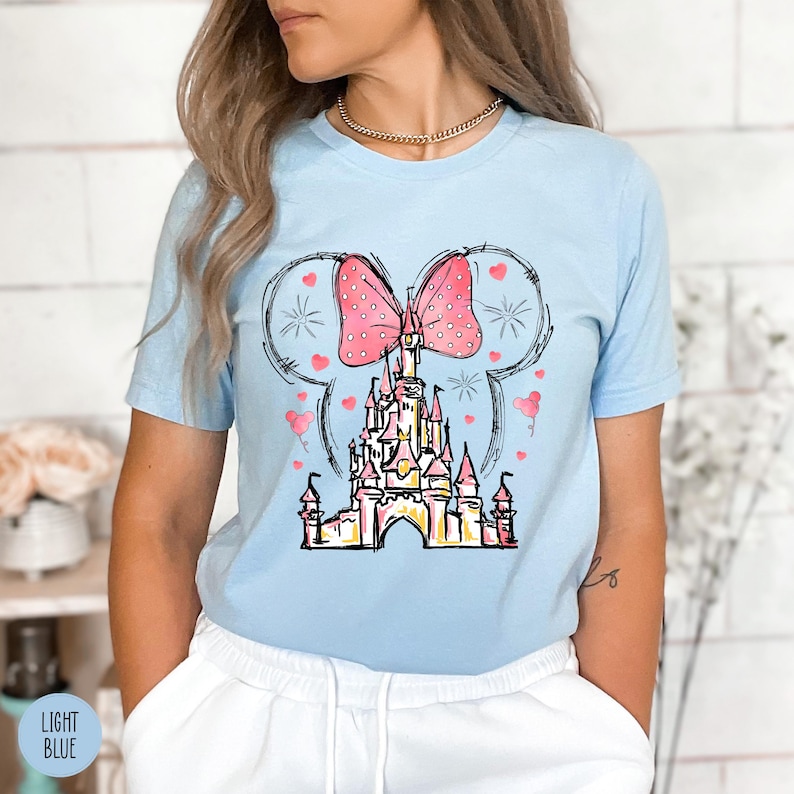 Puede incluir: Camiseta azul claro con un dise&ntilde;o de castillo, un lazo rosa y detalles de coraz&oacute;n. El dise&ntilde;o est&aacute; enmarcado por un contorno de oreja de Mickey Mouse. Las palabras "LIGHT BLUE" est&aacute;n impresas en la parte inferior izquierda de la imagen.