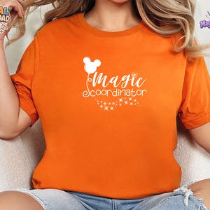 May include: A bright orange short-sleeved t-shirt featuring a white graphic print on the chest. The design includes a stylized mouse-ear balloon, the text "Magic coordinator" in a cursive font, and a trail of small sparkling stars below.