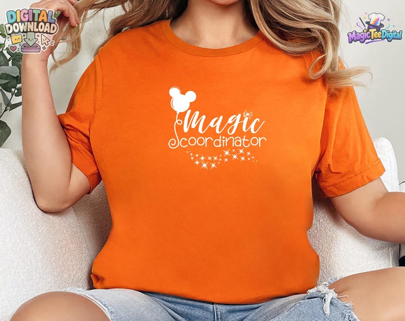 May include: A bright orange short-sleeved t-shirt featuring a white graphic print on the chest. The design includes a stylized mouse-ear balloon, the text "Magic coordinator" in a cursive font, and a trail of small sparkling stars below.